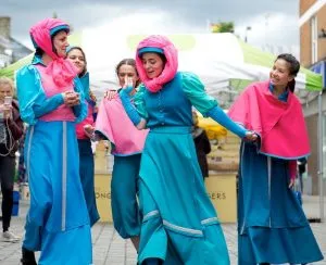 A group of women in blue & pink late 18th century long skirts & puffed sleeves walk down the road